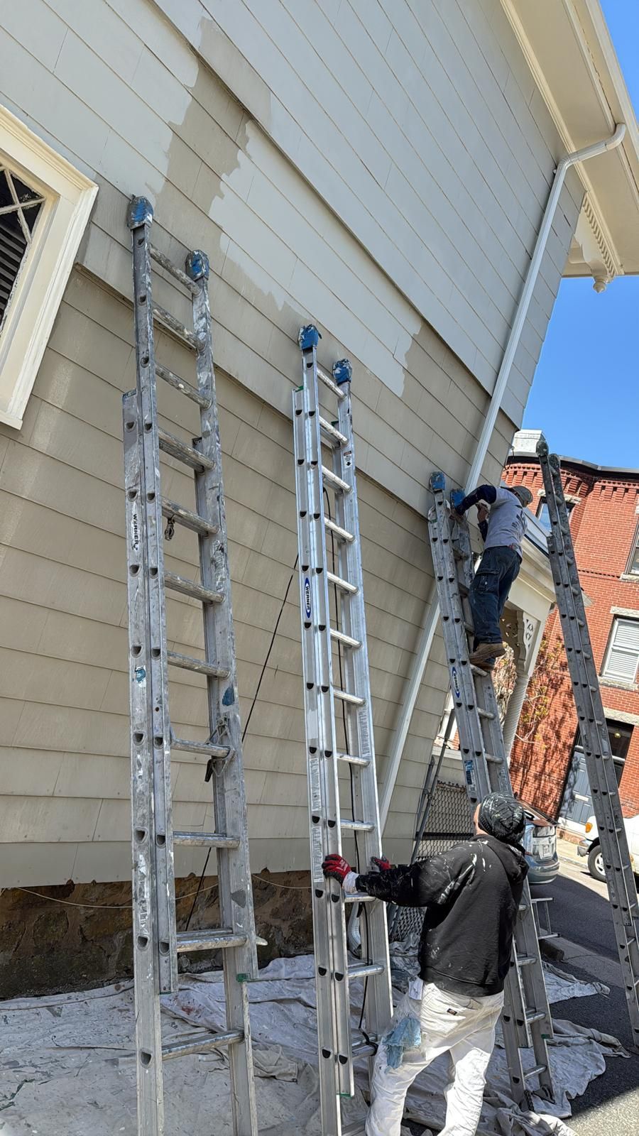 Two workers on ladders painting the exterior of a house beside a brick wall
