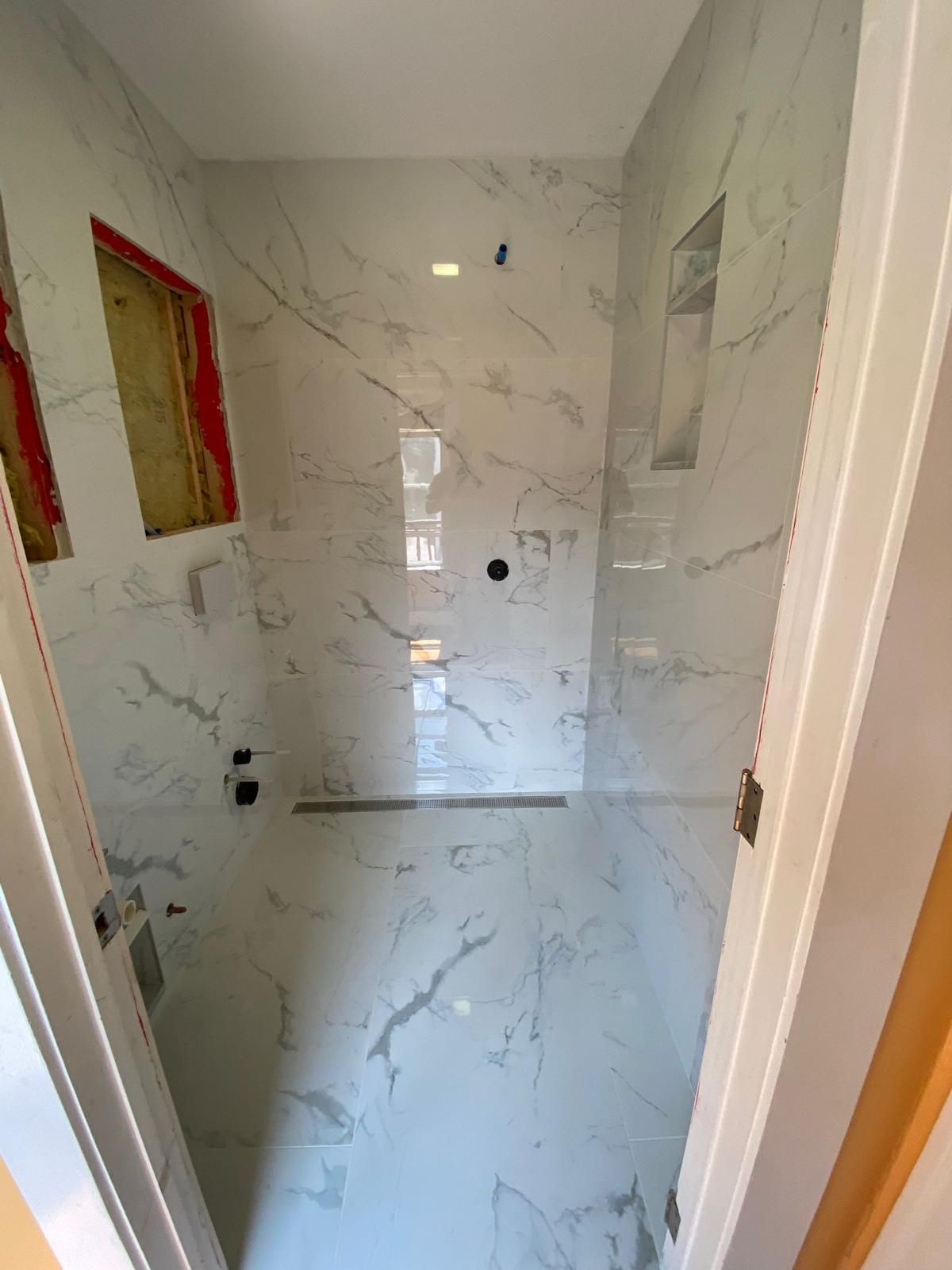 Narrow tiled shower with white marble-look walls, recessed niches, and unfinished openings for fixtures