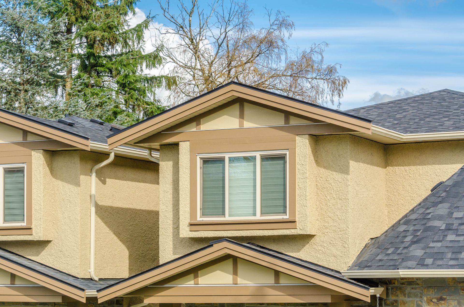 Two-story beige stucco townhouse with gray shingle roofs and trees in the background