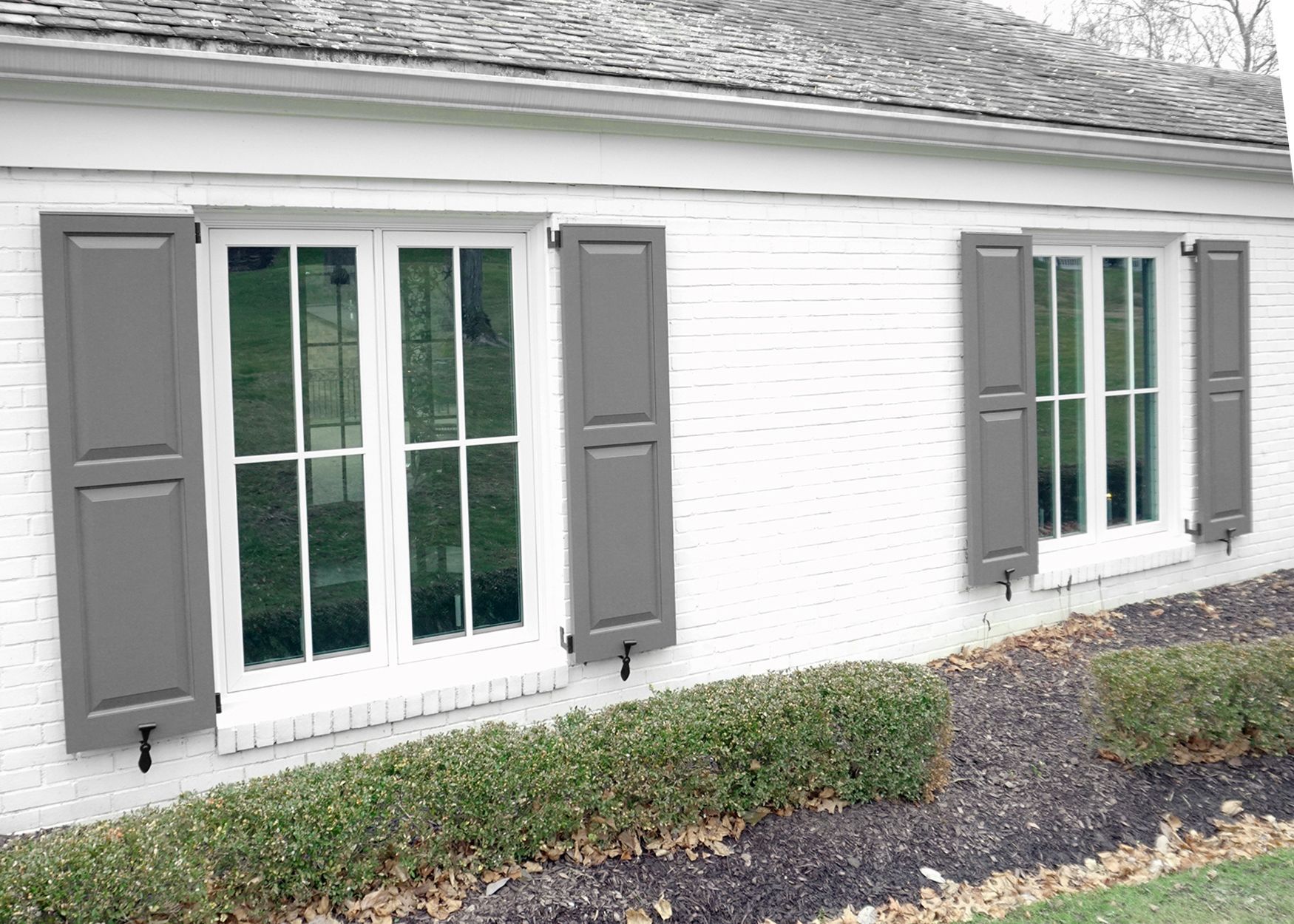 Two white-framed windows with gray shutters on a white brick building with a low hedge.
