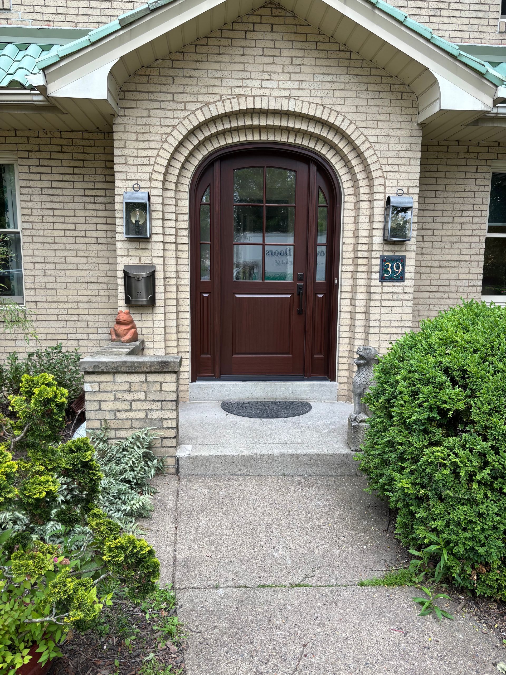 Brown arched front door, light brick facade, walkway, and green bushes.