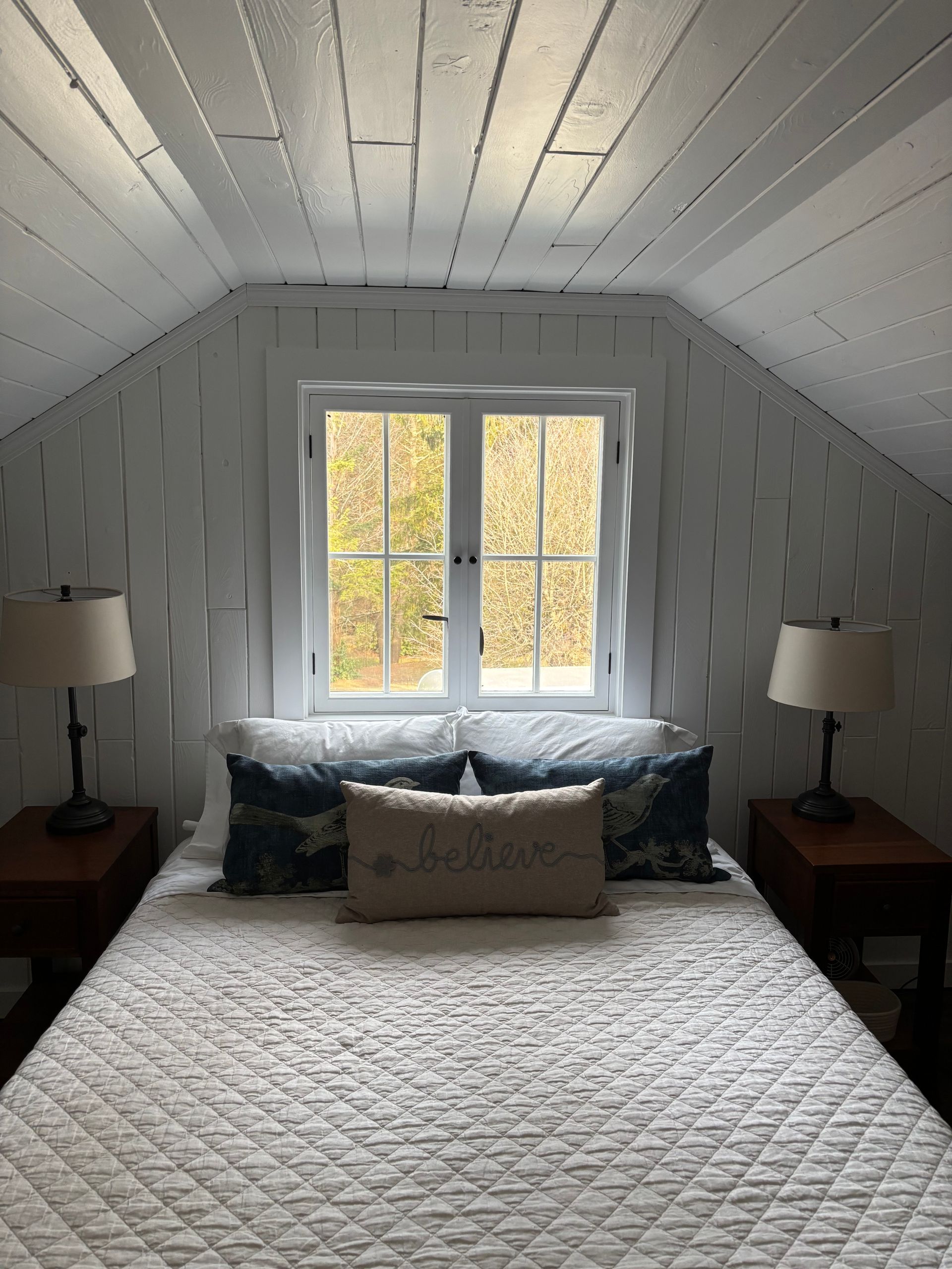 Bedroom with white walls, sloped ceiling, and a window overlooking a wooded area. Bed has pillows and two lamps.