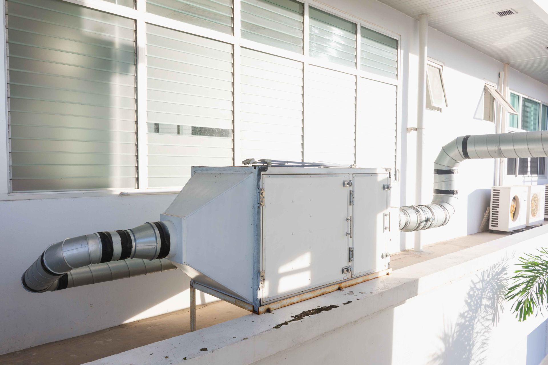 White ventilation unit with silver ductwork on a building exterior, next to windows.