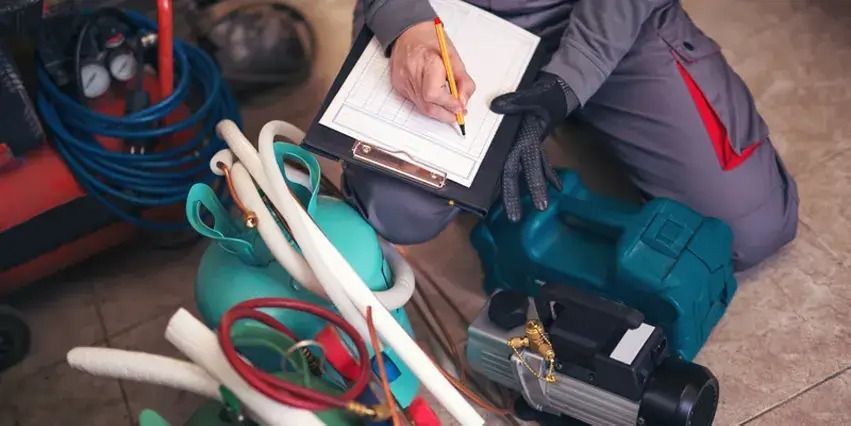 Person in grey overalls writing on a clipboard, surrounded by equipment, in a work setting.