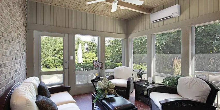 Sunroom with white sofas, brick wall, large windows, and an air conditioner.