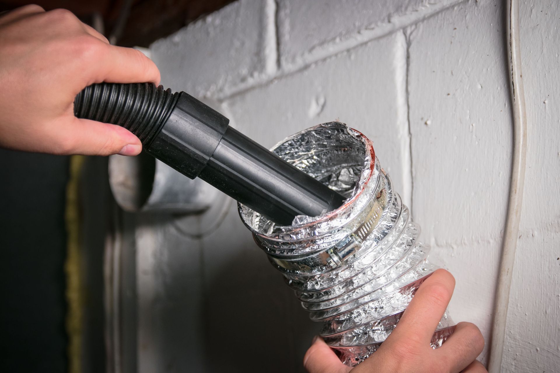 Person cleaning dryer vent with a vacuum hose. Silver vent duct against white wall.