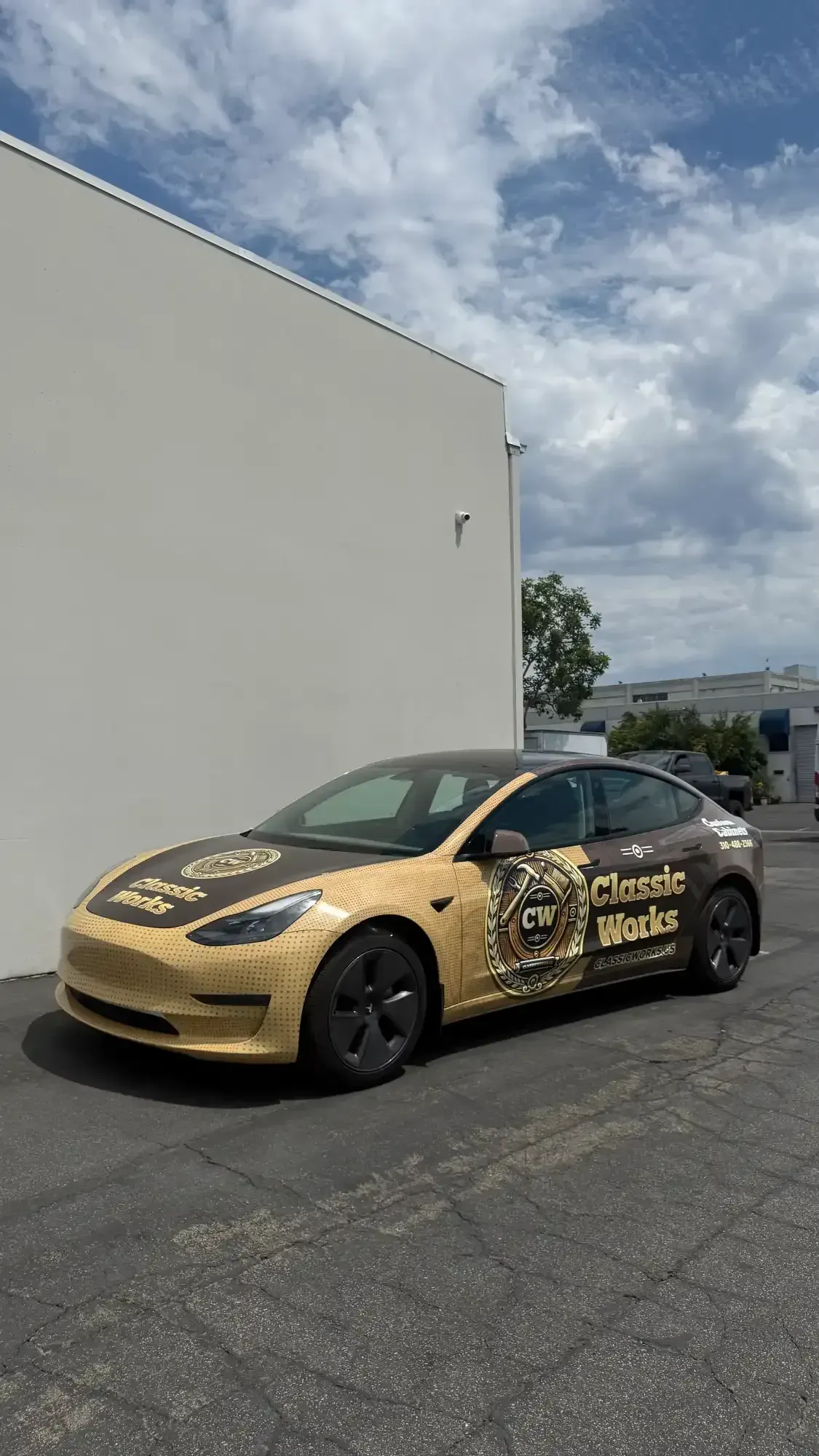 Gold-wrapped Tesla car parked in front of a white building with advertising 