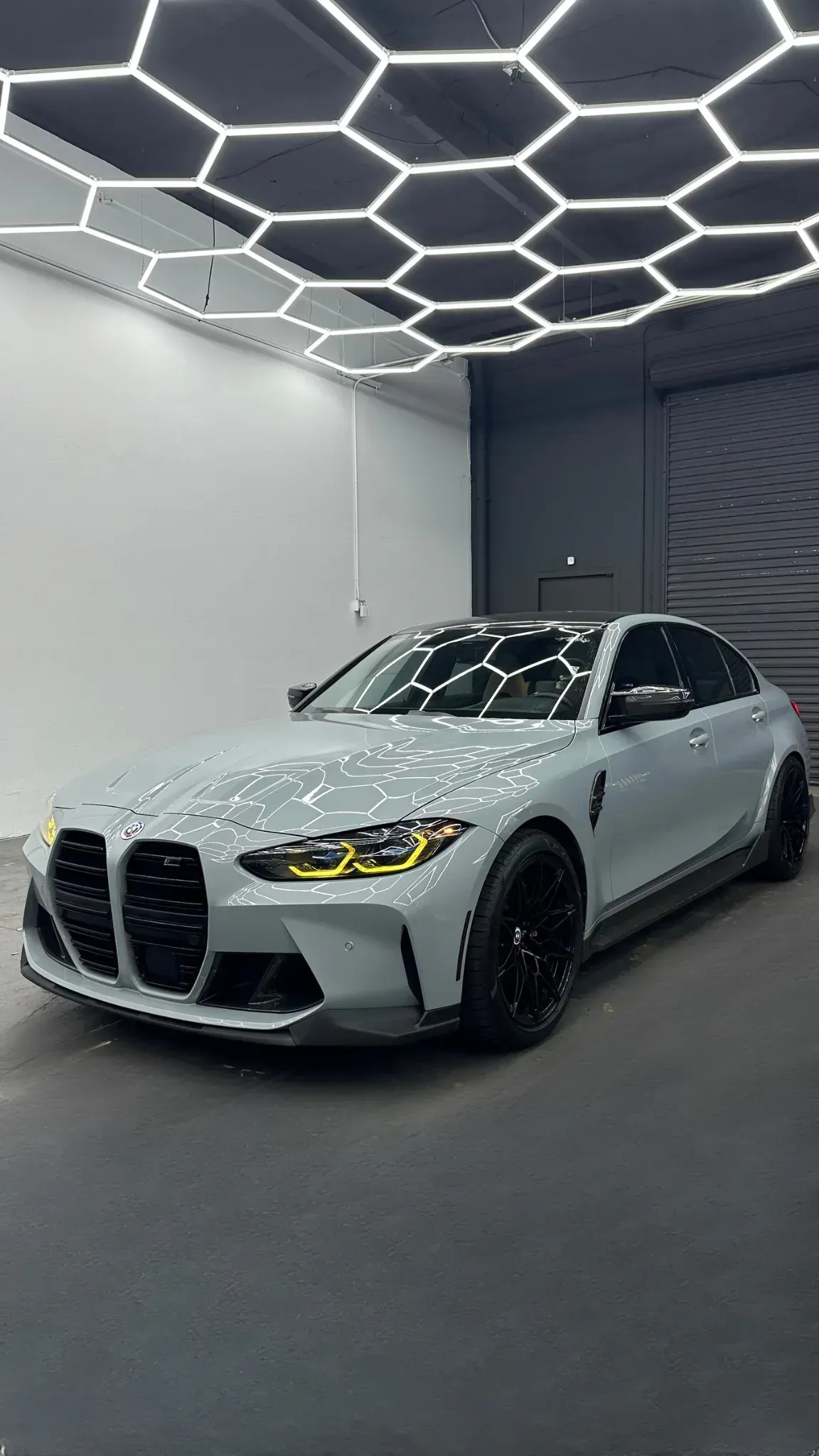 Gray BMW M4 coupe under hexagon ceiling lights in a garage.