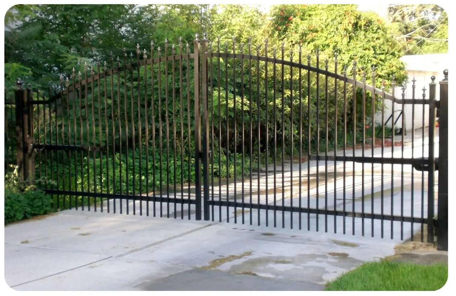 Black wrought iron gate on a concrete driveway, leading to greenery.