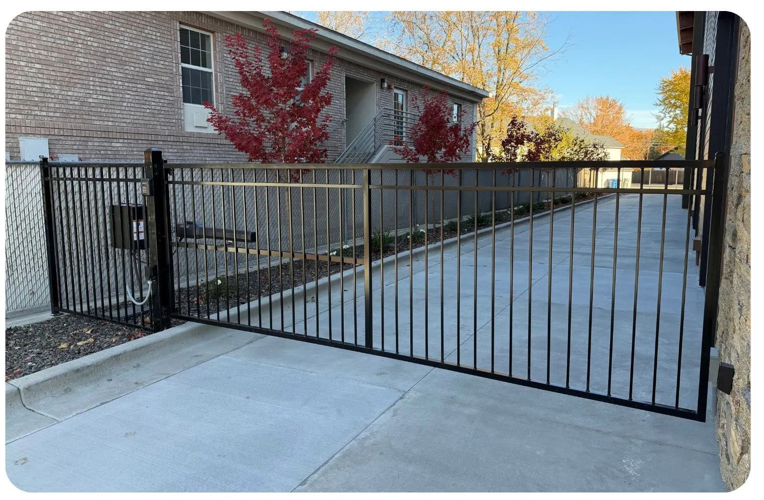 Black metal fence encloses a concrete driveway beside a building with red vines.