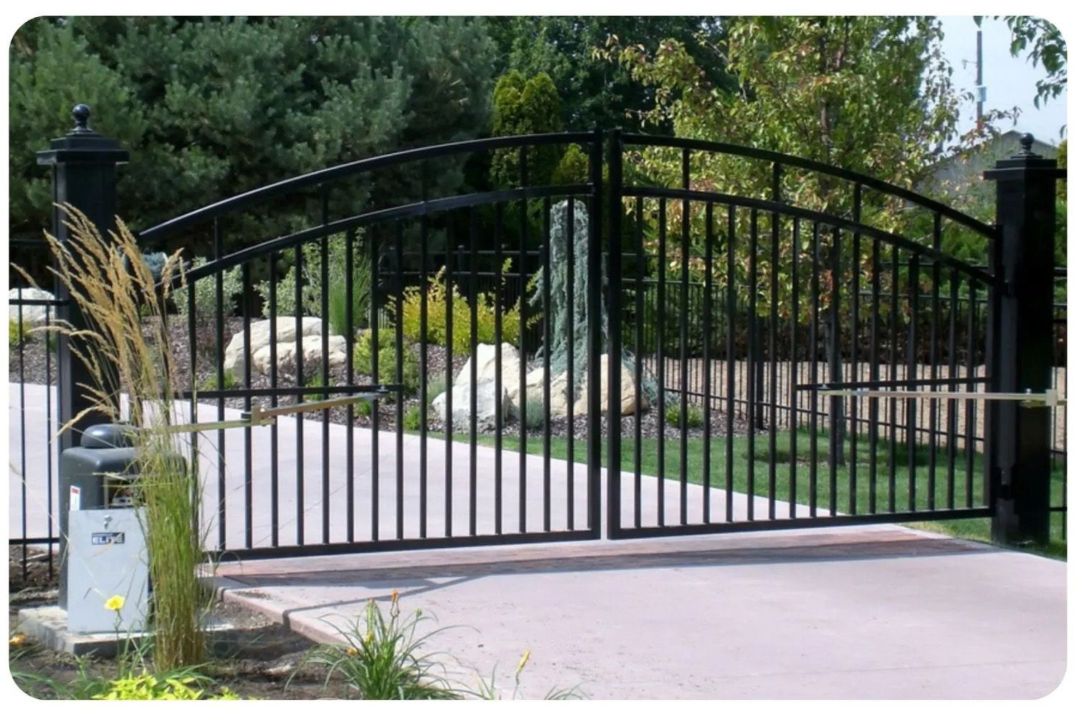 Black wrought-iron driveway gate, arched design, on concrete, leading to a landscaped yard.
