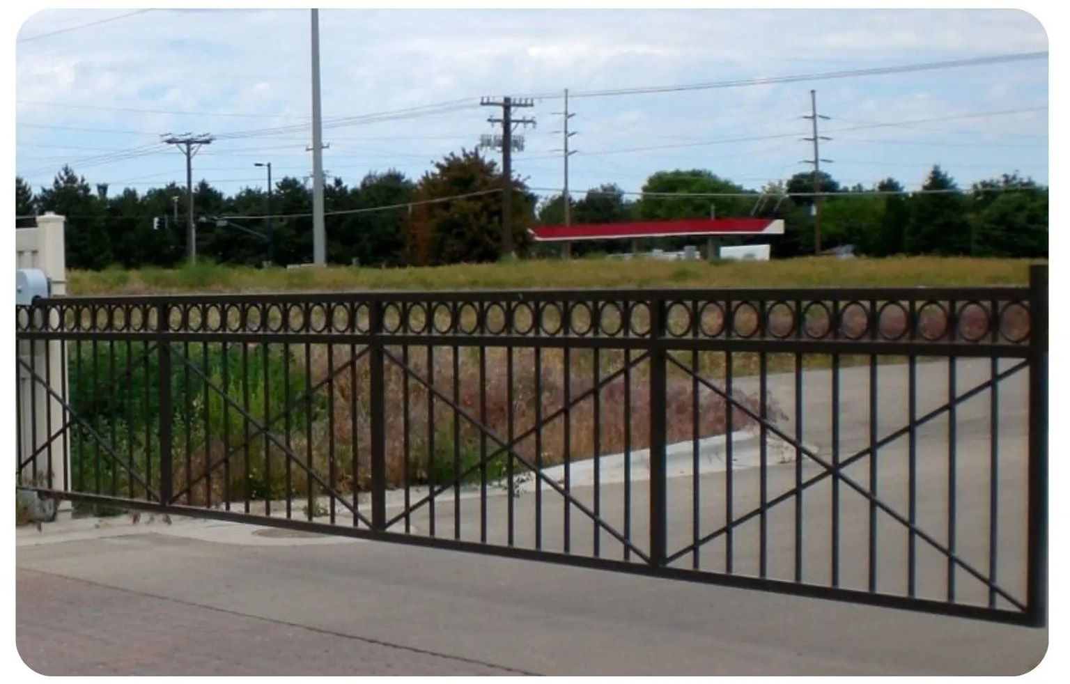 Black metal fence with decorative top, in front of a grassy field, with a building and power lines in the distance.