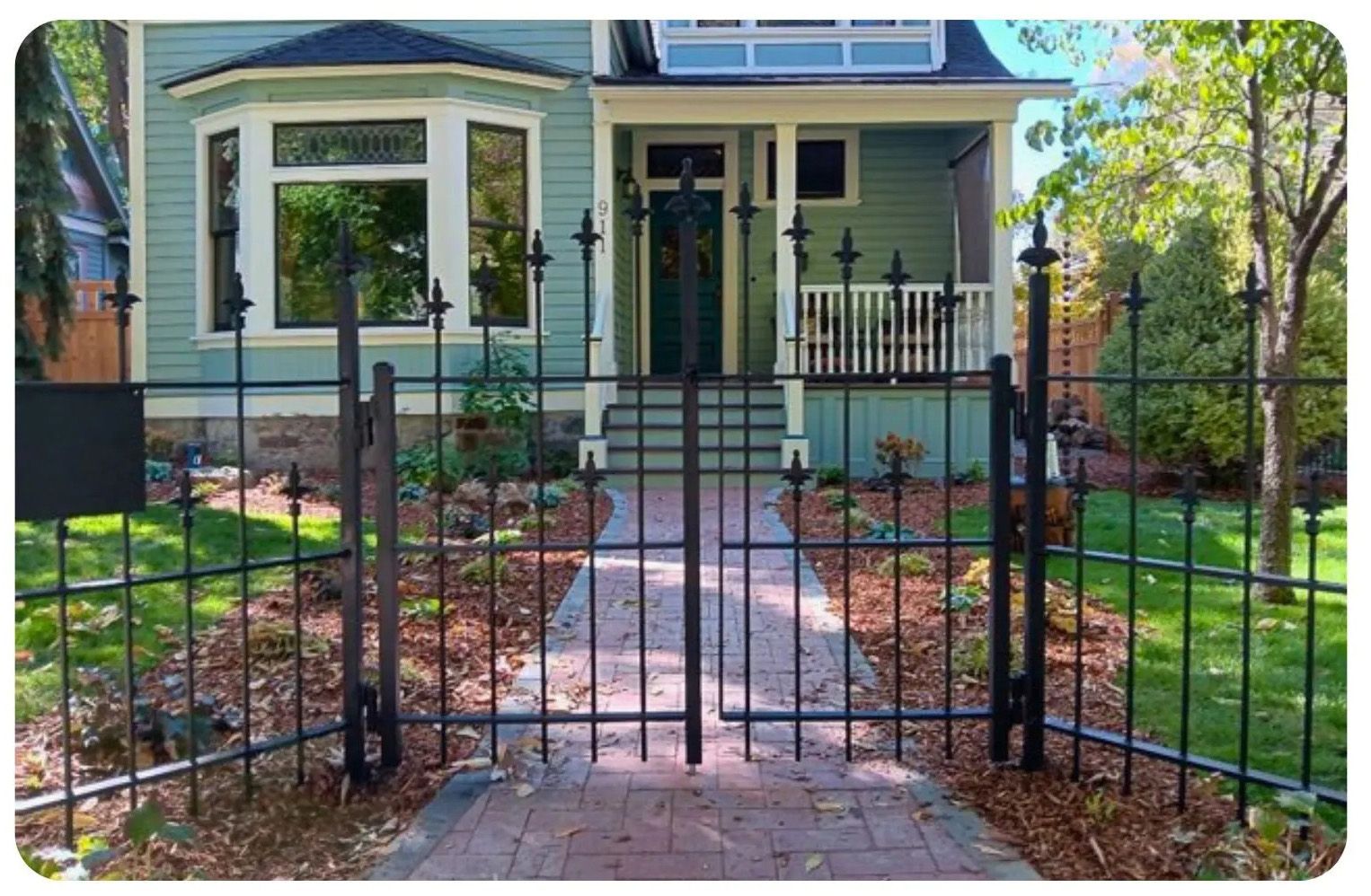 Black iron fence and gate leading to a light-green house with a porch and brick walkway.