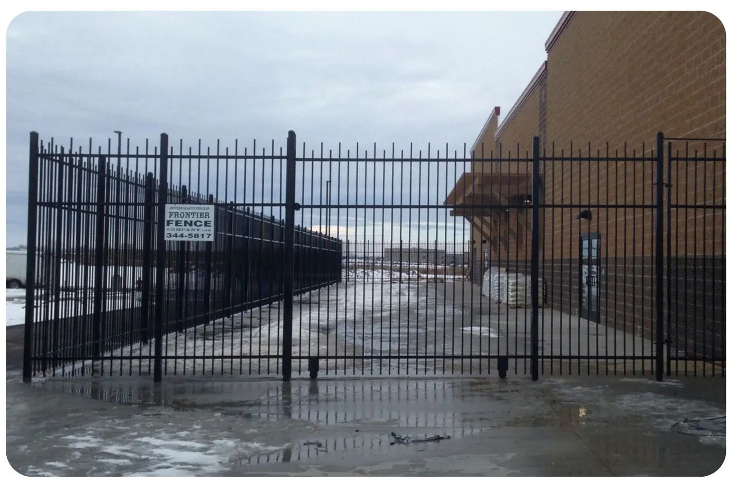 Black metal fence blocking a wet, concrete area next to a brick building. Sky is overcast.