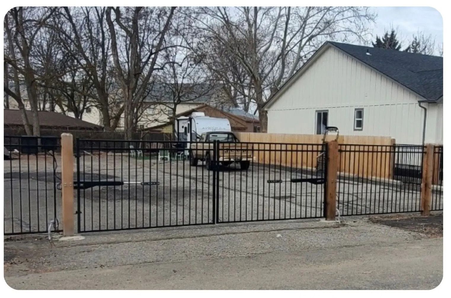 Black metal gate in front of gravel lot, with a house and vehicle visible.