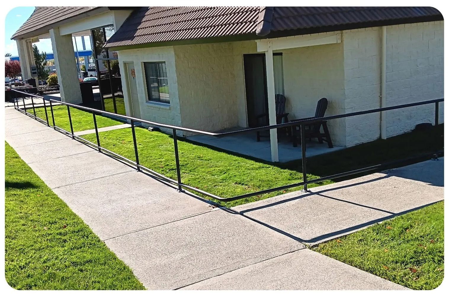 Exterior view: Concrete walkway with black railing alongside a building with a brown roof and small lawn.