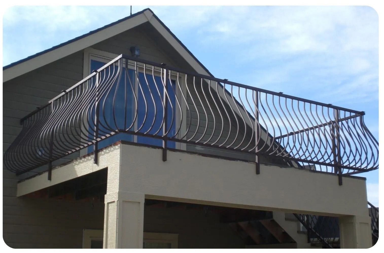 Black wrought-iron railing on a balcony, against a house and blue sky.