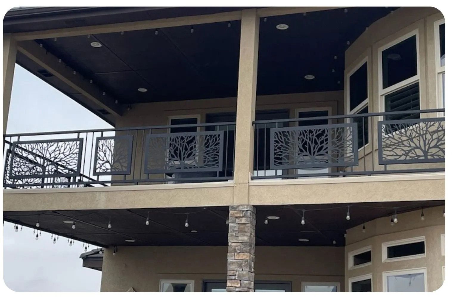 Two-story house with a balcony featuring tree-design metal railings, string lights, and a tan exterior.
