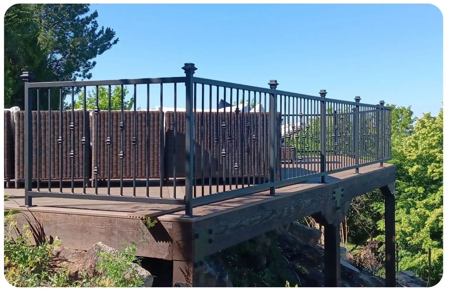 A metal railing surrounds an outdoor viewing platform with wooden backing; trees and blue sky visible.