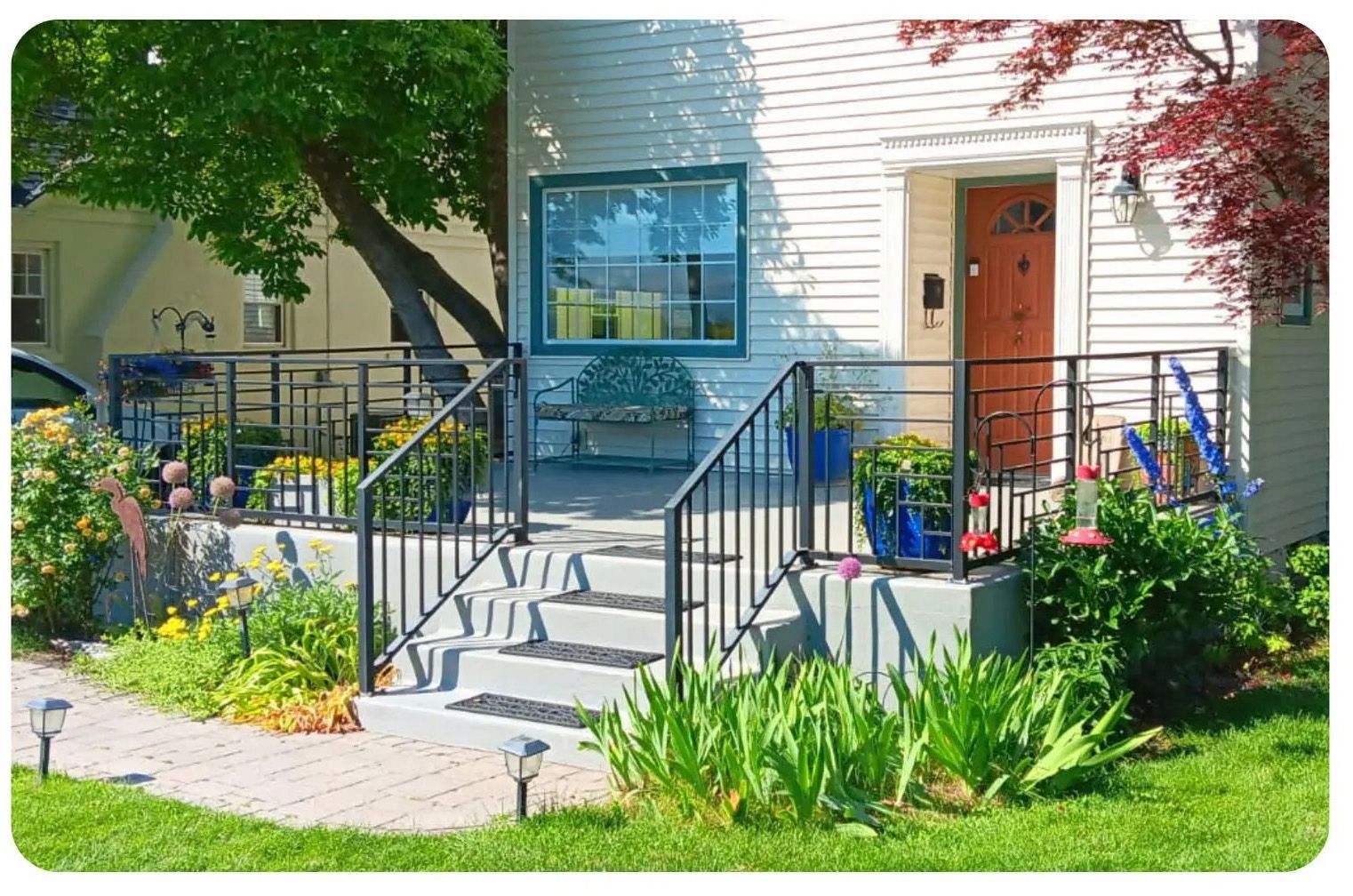 A white house with steps and a porch, featuring a black railing, garden and a path.