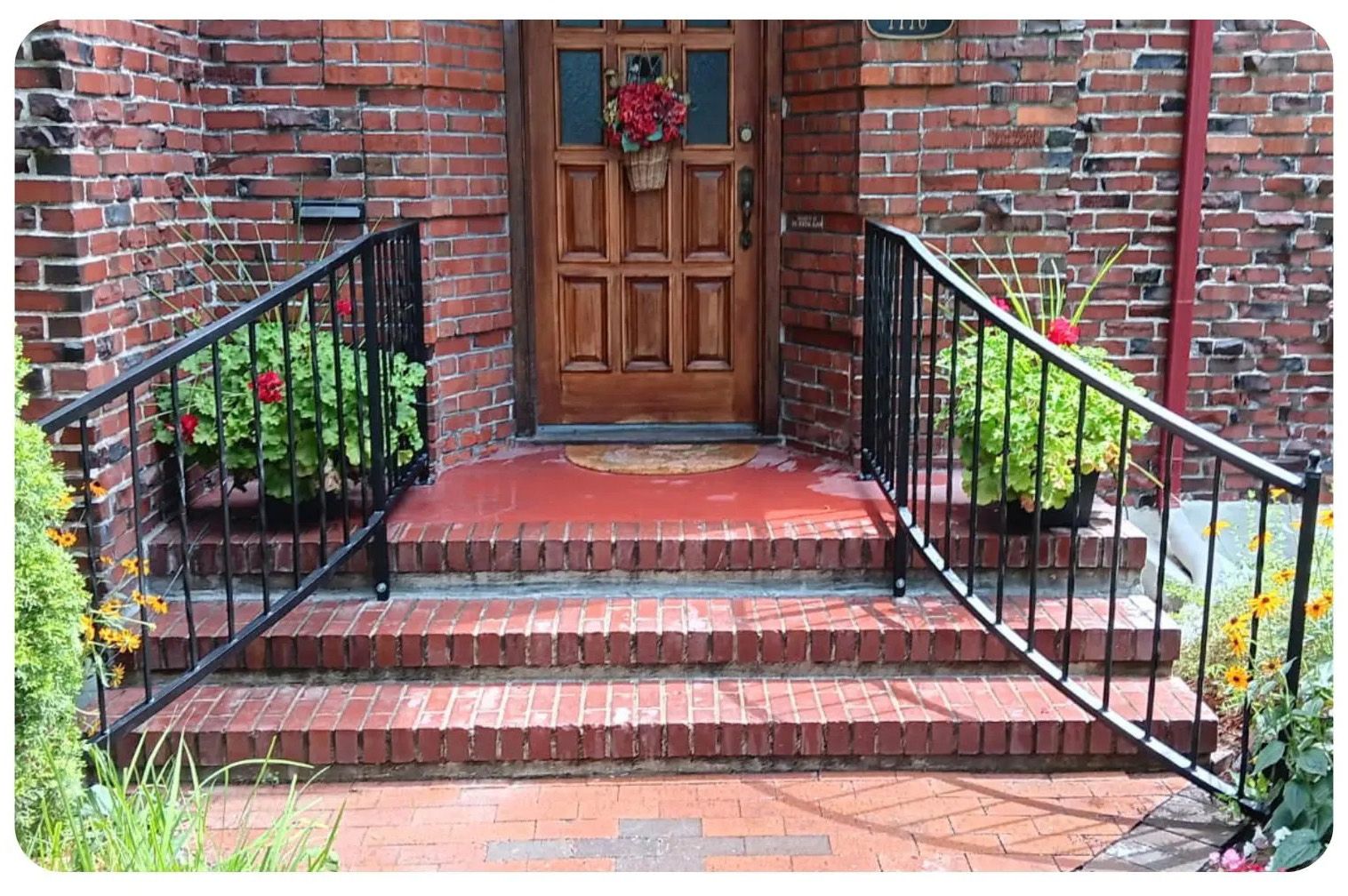 Brick steps leading to a wooden door, flanked by black iron railings and potted plants.