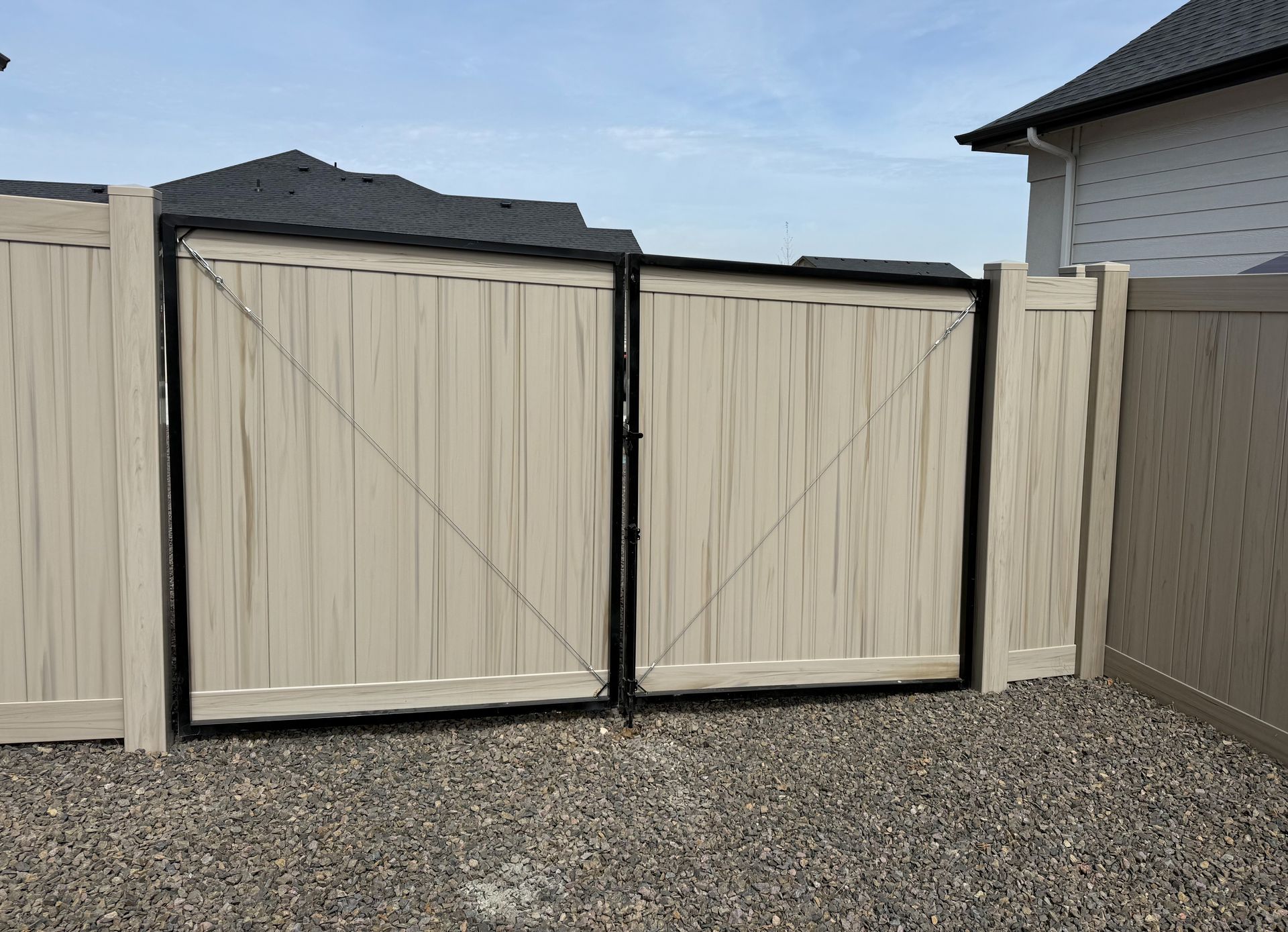 Double-gate entrance with light-colored panels and black metal framing, situated between vinyl fence sections over gravel.