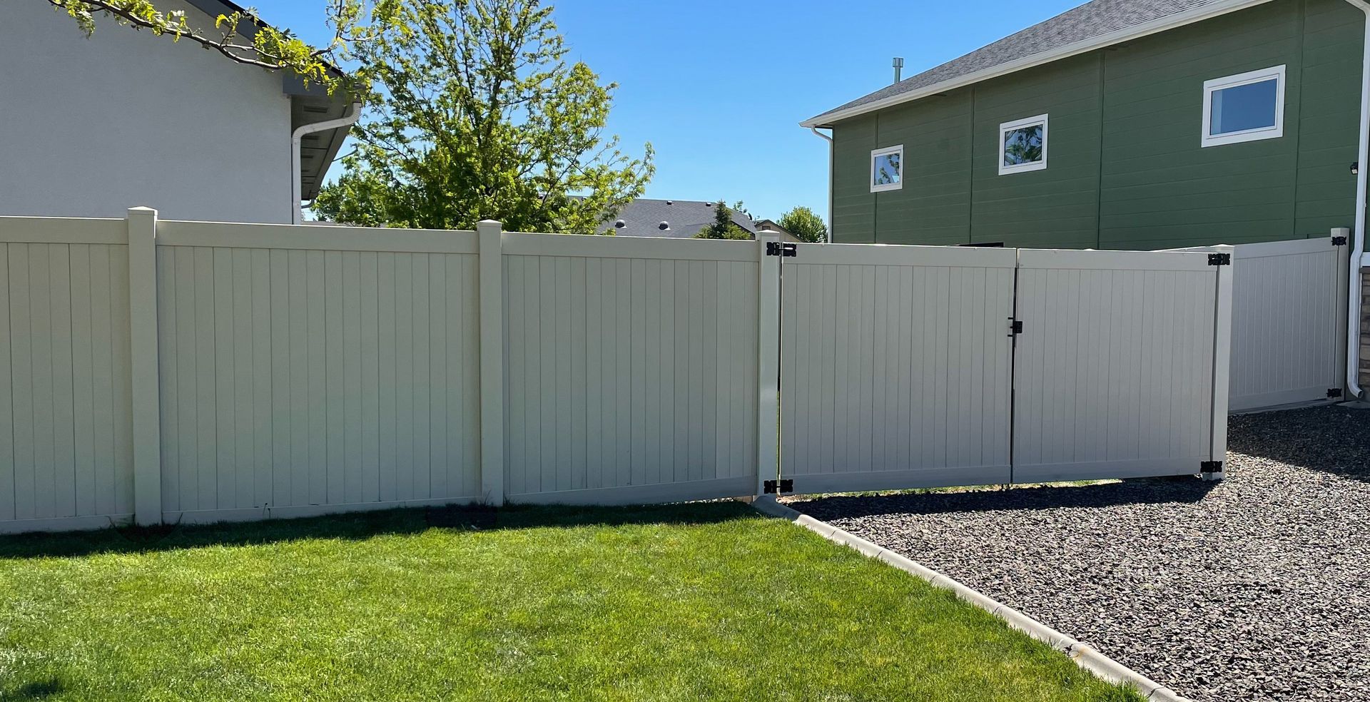 A white vinyl fence with a double gate stands between a grassy lawn and a gravel driveway on a sunny day.