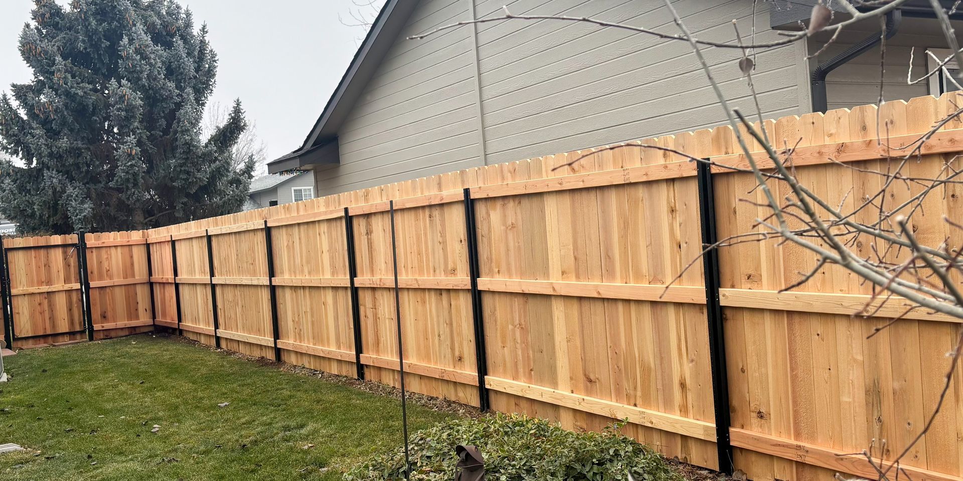 A long, new wooden privacy fence with black metal support posts borders a grassy yard next to a house.