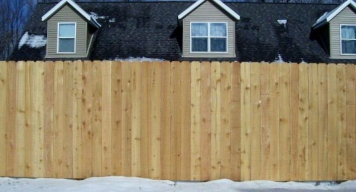 A light wood privacy fence stands in front of a house with three dormer windows and a dark shingled roof.