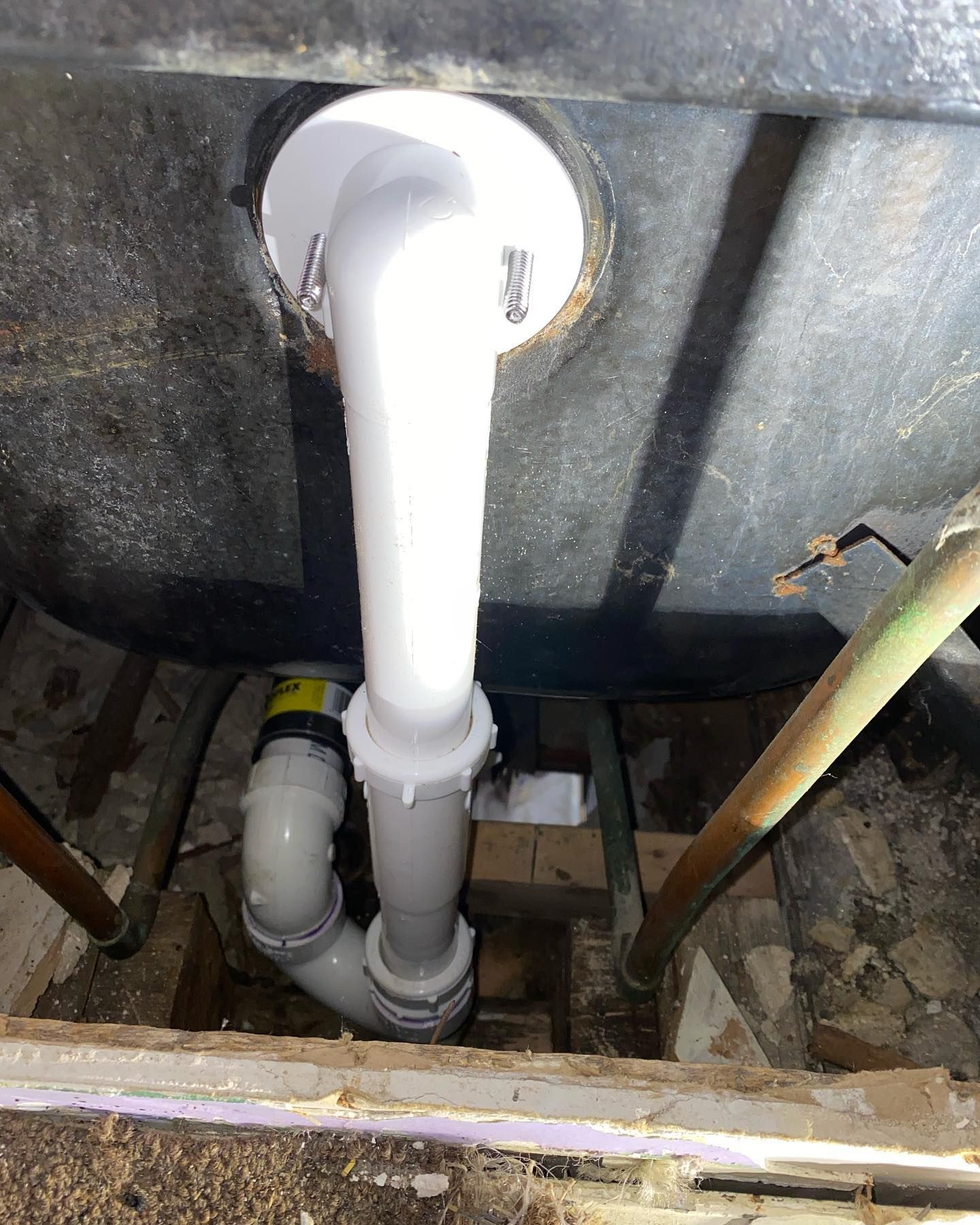 Underside view of a bathtub drainpipe, showing white PVC pipes in a small, dark space between wood beams.
