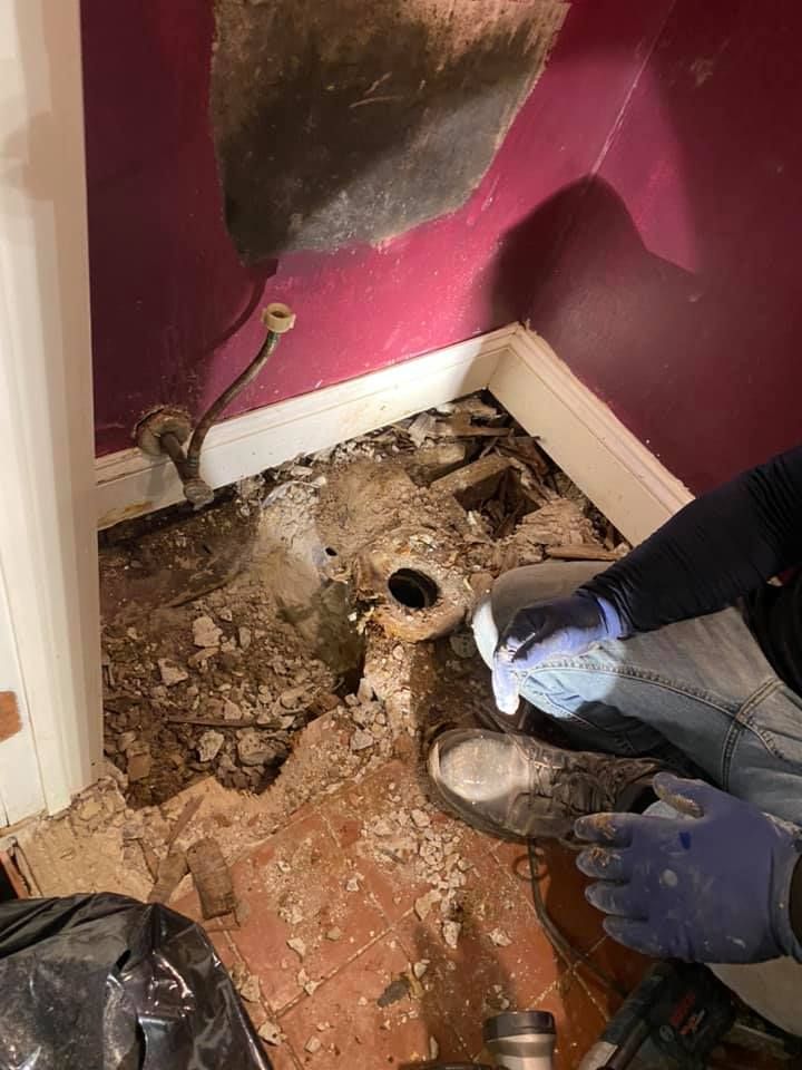 Person in blue gloves examines exposed plumbing and floor in a bathroom with damaged walls.