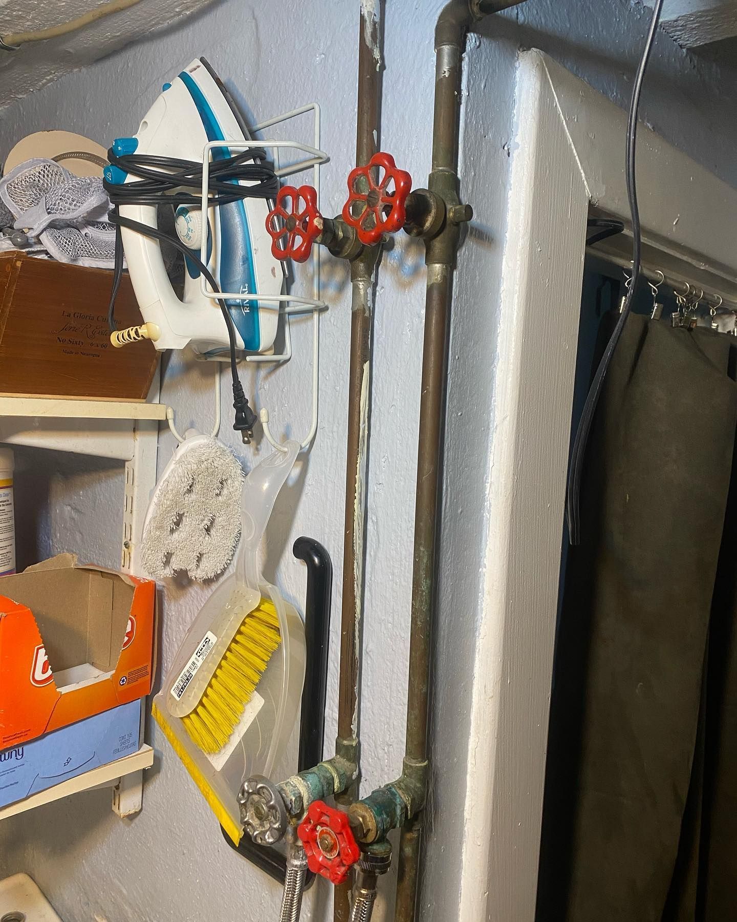 Pipes with red and silver valves on a wall, next to a broom, sponge, iron, and shelves.