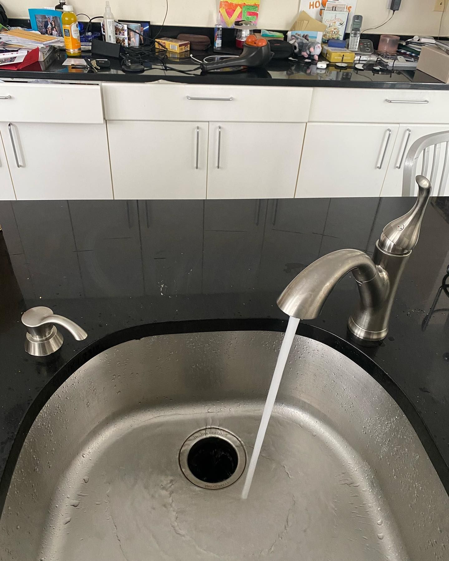 Stainless steel kitchen sink with water running, faucet and soap dispenser, black countertop, white cabinets.