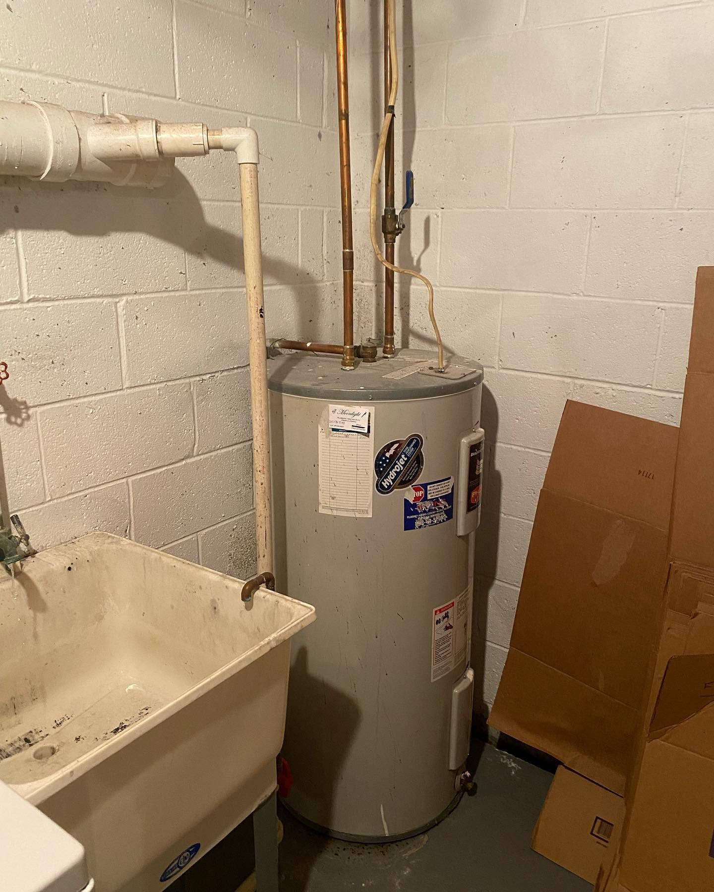 A gray water heater in a basement laundry area with a utility sink and cardboard boxes.