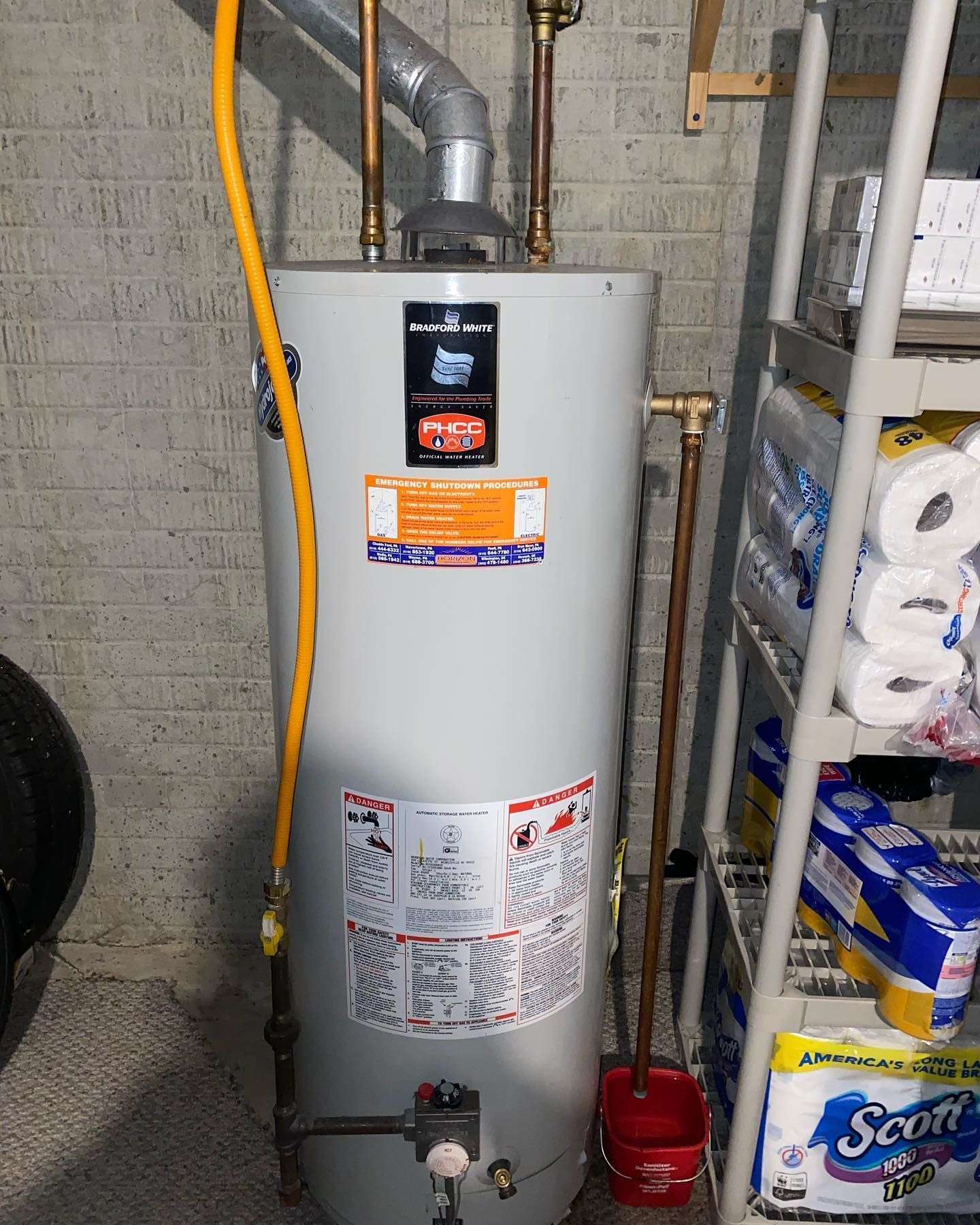A vertical water heater in a basement next to a shelf of supplies, with copper pipes and a yellow gas line.