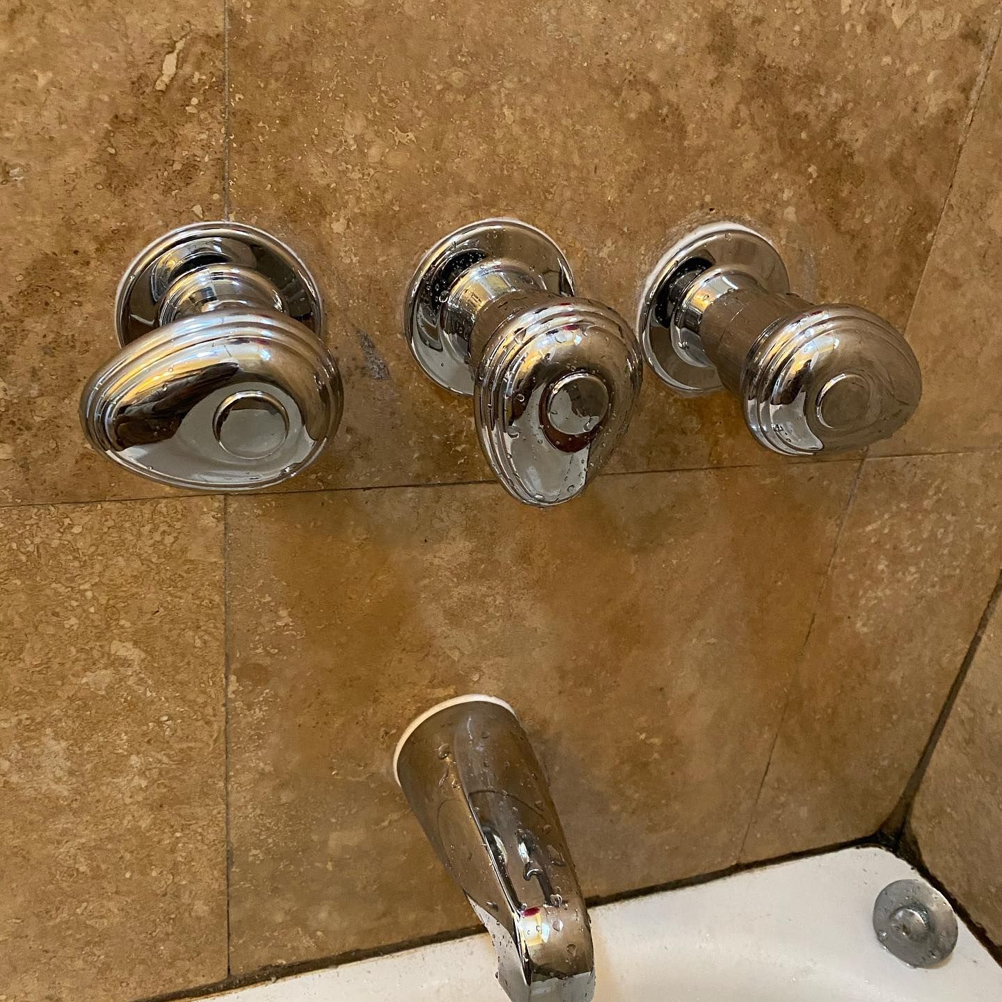 Shiny chrome bathroom faucet and three matching knobs against beige tile.
