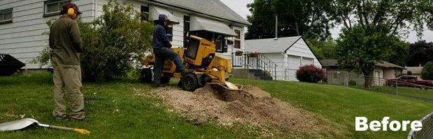 Worker on a yellow skid steer removing a dirt mound beside a house and driveway, with another person nearby.
