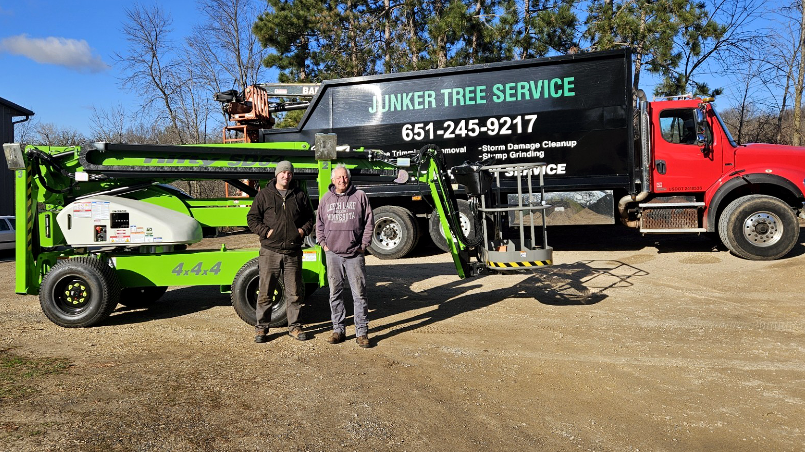 Tree service truck and lift on a gravel lot with two workers standing beside it.