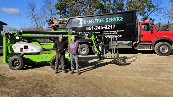 Tree service truck and lift on a gravel lot with two workers standing beside it.