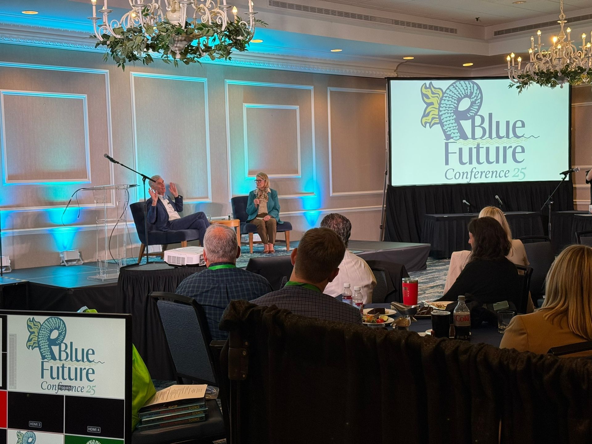 A conference stage with two speakers in chairs. A screen displays a 