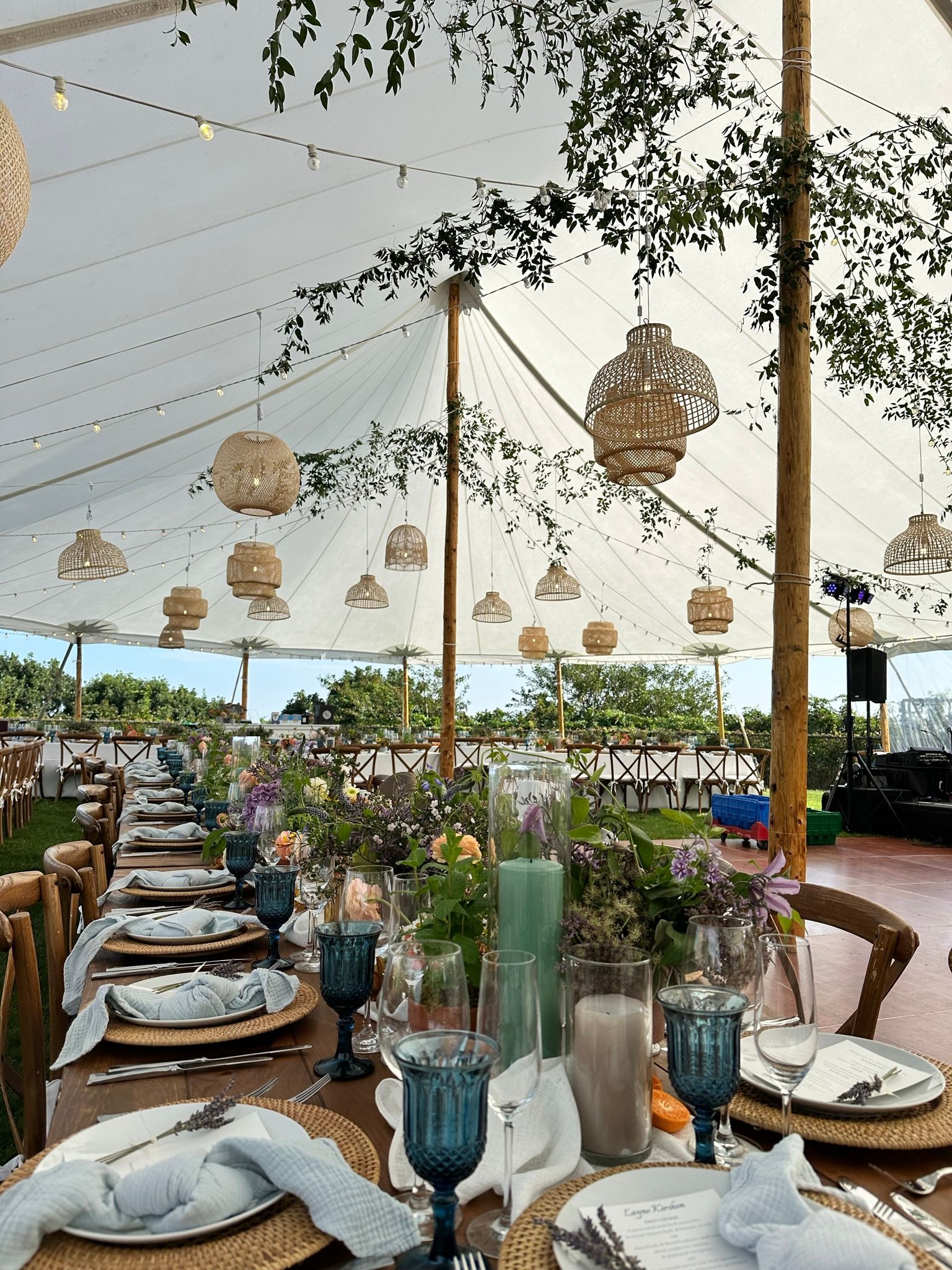 Long table set for an outdoor event under a tent, with floral arrangements and hanging lanterns.