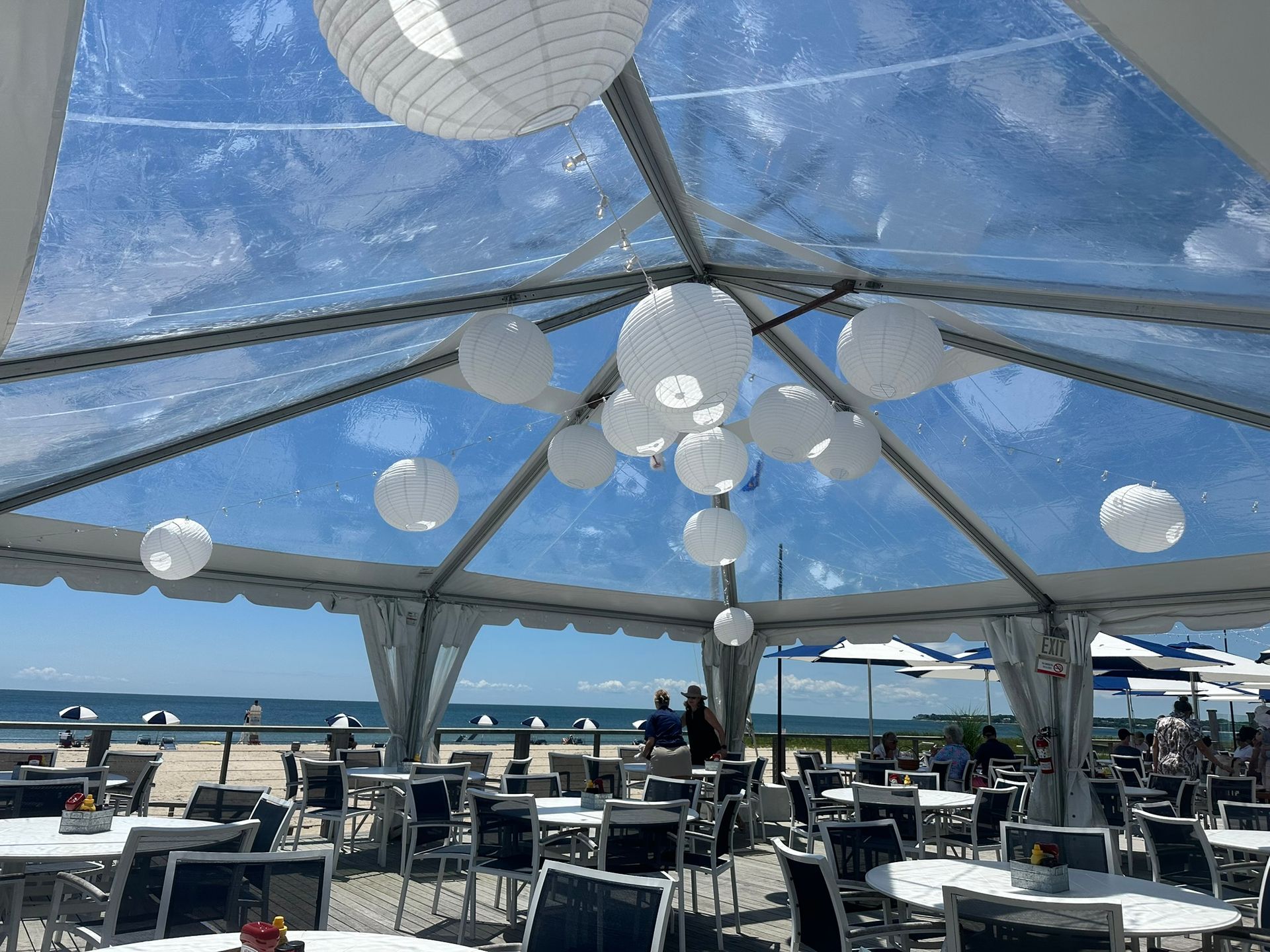 Beachside restaurant under a clear tent, decorated with paper lanterns, tables, and people enjoying the view.