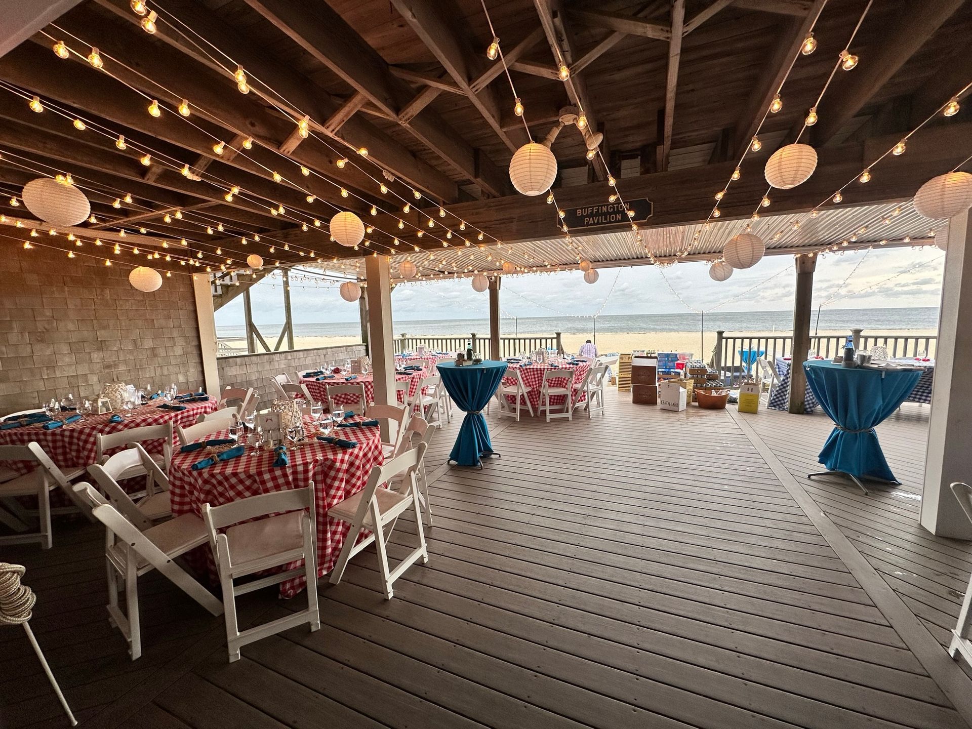 Outdoor event space with tables set for a meal, string lights, paper lanterns, and beach view.