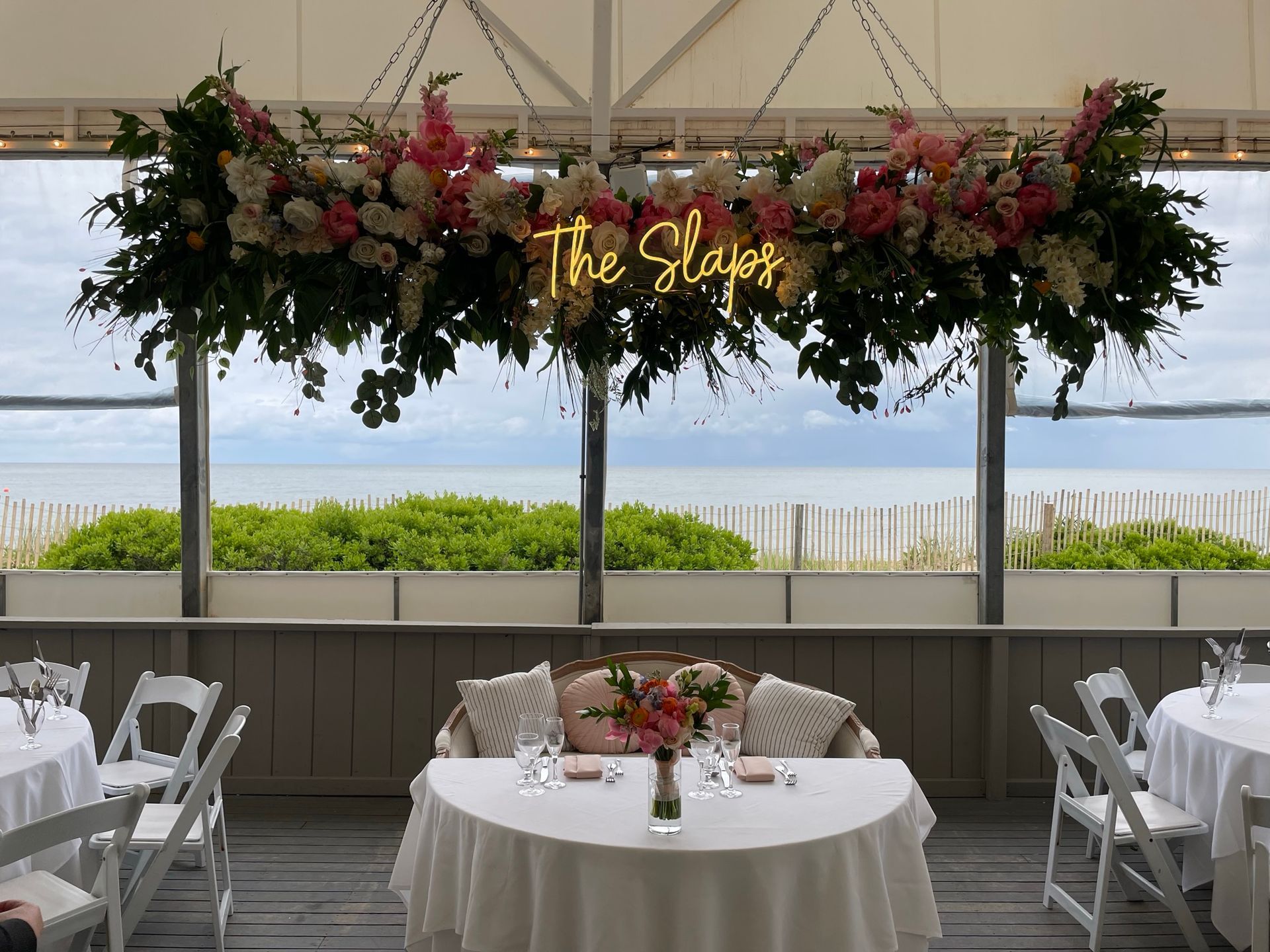Wedding reception decor with floral chandelier, table setting, and ocean view.