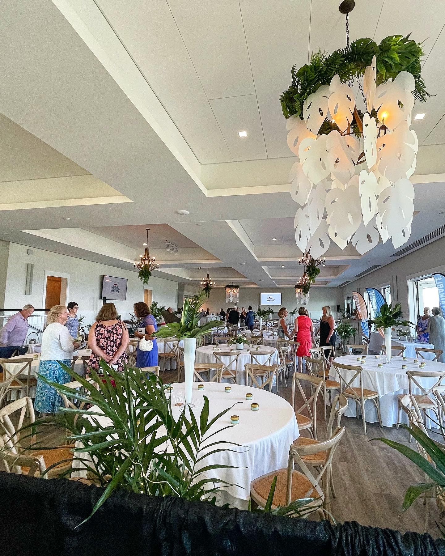 Elegant event space, tables set, people mingling. White linens, greenery decor, unique chandelier.
