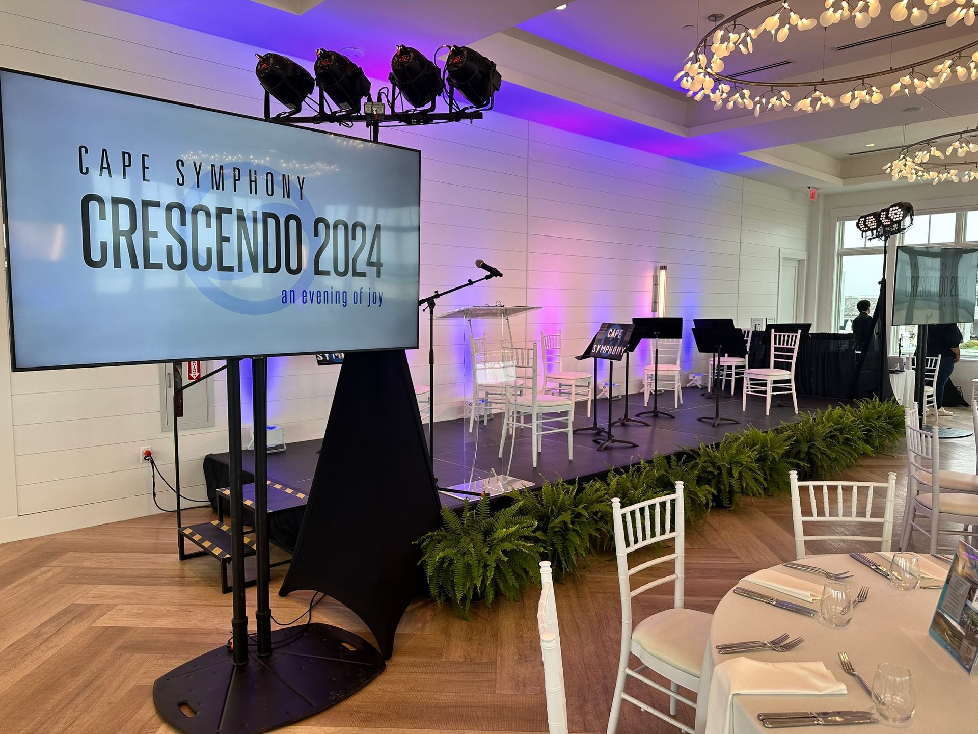 Stage set for Crescendo 2024 event, with a screen, lighting, and seating. White chairs and round tables are set.