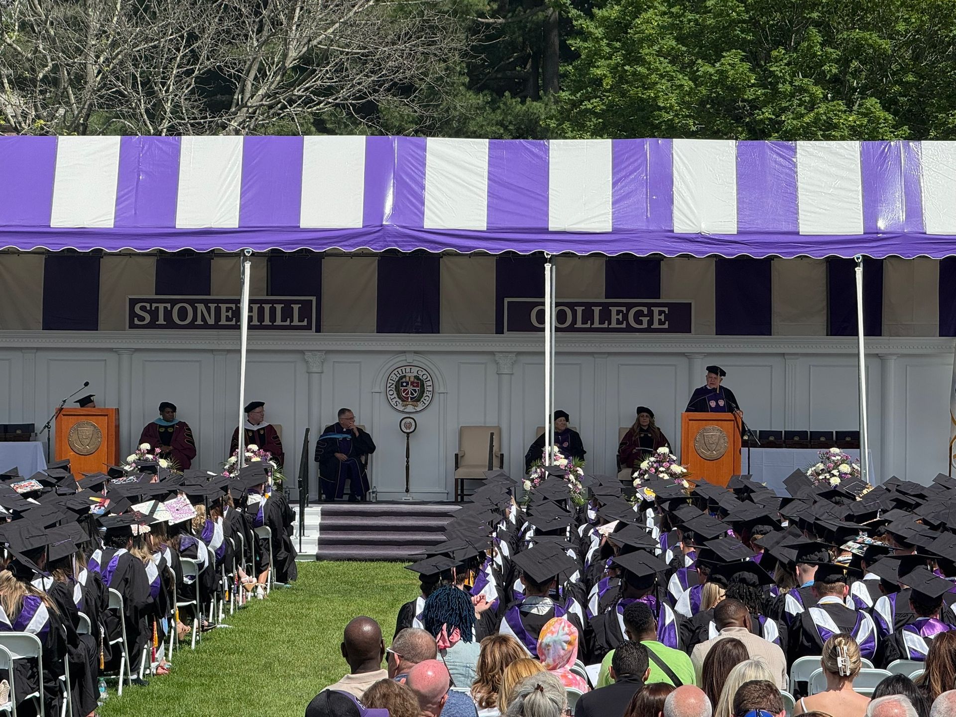 Stonehill College graduation ceremony under a purple and white tent. Graduates in caps and gowns face the stage.
