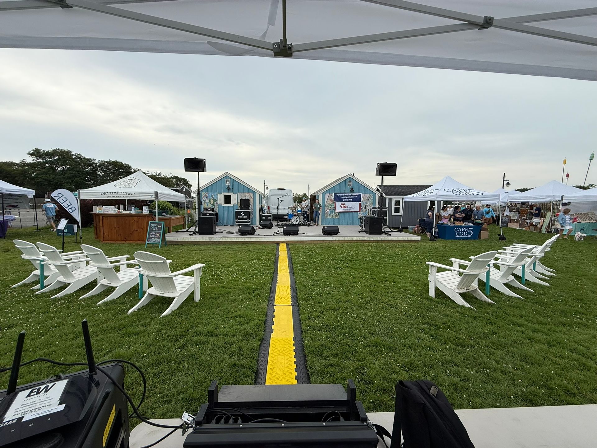 Outdoor stage set up with two small blue houses, chairs, and vendors under tents on grass.