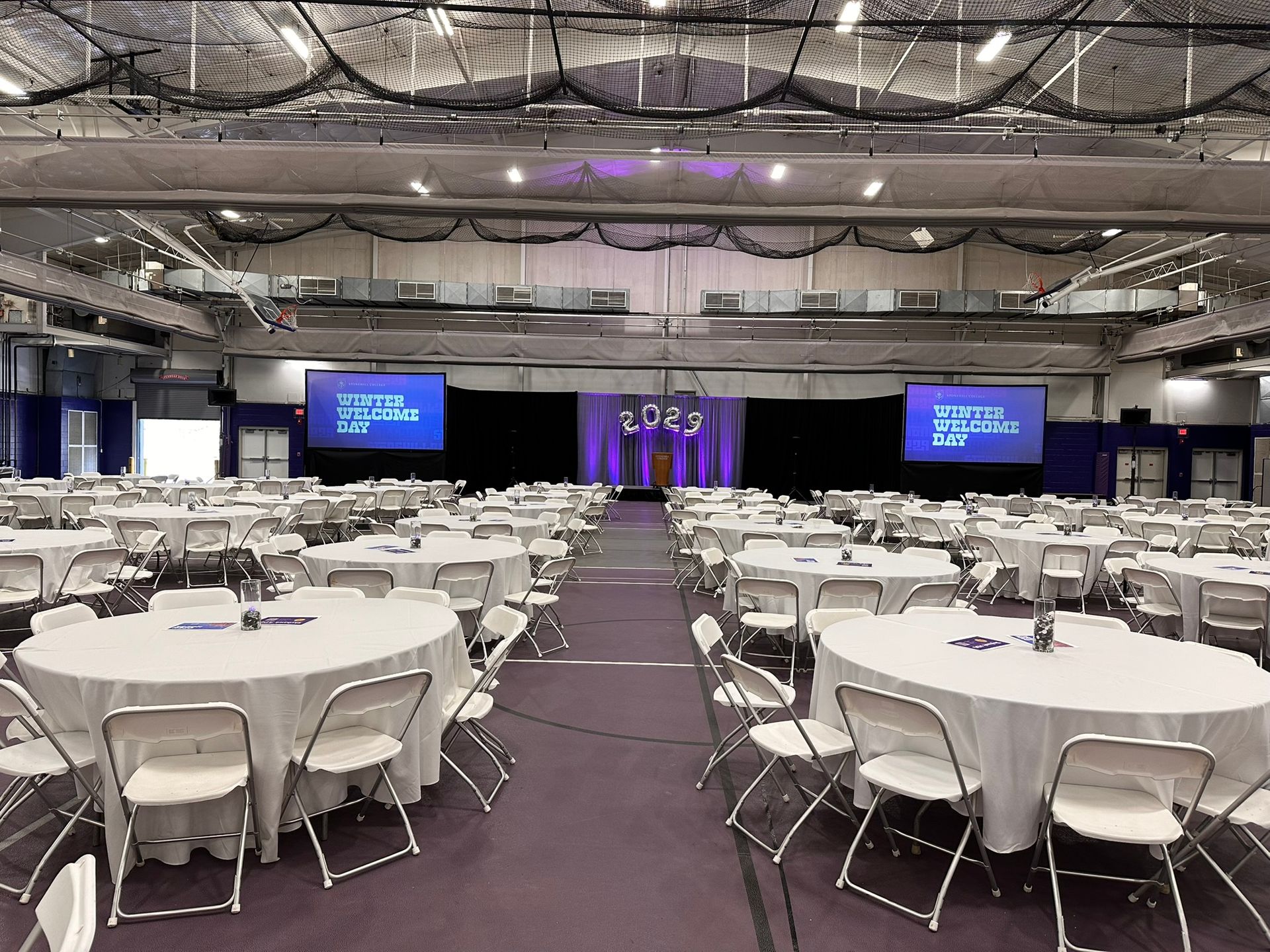 Conference hall set for an event with round tables covered in white linens and white chairs. Blue screens and stage in the back.