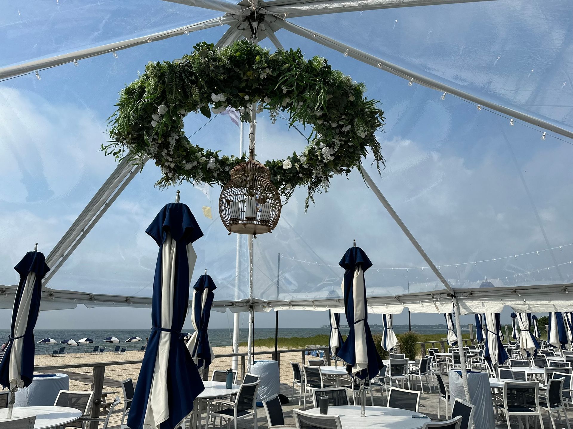 Clear tent interior overlooking a beach. A floral wreath and chandelier hang from the ceiling. Blue and white umbrellas are closed.